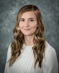 A woman insurance group company with long wavy light brown hair wearing a light-colored knit sweater, posed in front of a gray mottled background.
