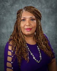 A woman from insurance group company with long, braided hair wearing a purple top with cutout sleeves and a silver necklace poses in front of a gray textured background.