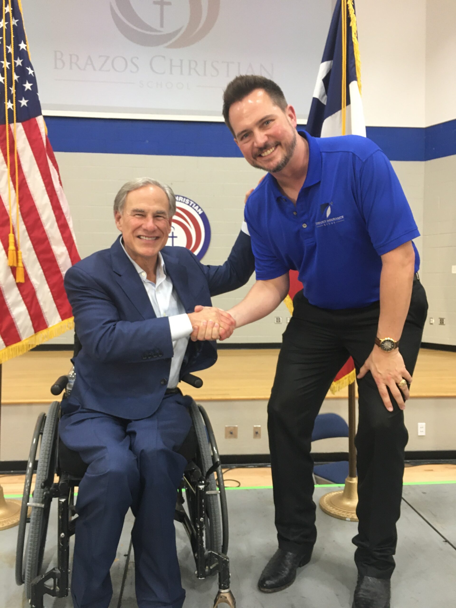 Two men smiling and shaking hands on a stage; one is seated in a wheelchair. American and Texas flags are visible in the background along with a Brazos Christian School sign.