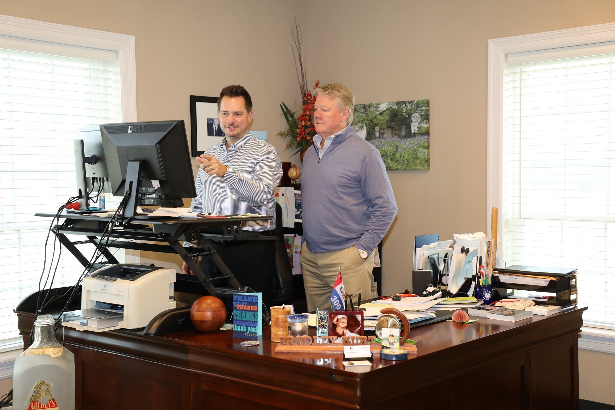 Two men stand at a desk with computer monitors in a well-lit office, surrounded by office supplies, personal items, and paperwork.