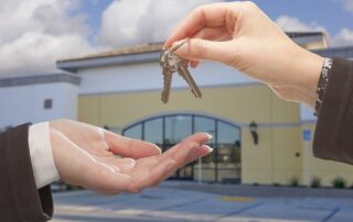 A person hands over a set of keys to another person in front of a modern commercial building.
