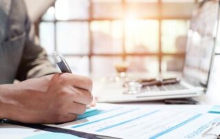 A person in business attire fills out a form at a desk with documents, a laptop, and a pen in hand.