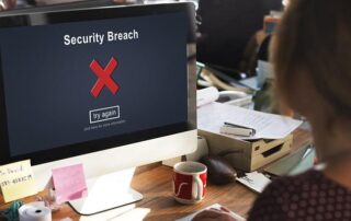 A computer screen displays a Security Breach message with a red X and try again button. A person is seated at the desk, facing the monitor.
