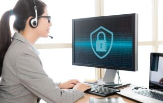 A woman wearing a headset and suit works at a desk with two computers, one displaying a cybersecurity lock icon on the monitor.