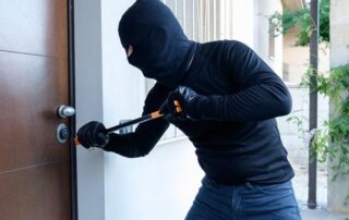 A person in a black mask and gloves uses a crowbar to pry open a door, appearing to attempt a break-in.