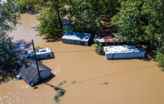 Aerial view of several houses partially submerged in floodwater, surrounded by trees, after severe flooding.