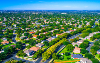 Aerial view of a suburban neighborhood with tree-lined streets, numerous single-family houses, and sprawling greenery under a clear blue sky.