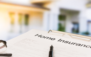 A document labeled Home Insurance on a clipboard, with a pair of glasses and a pen resting beside it, and a blurred house in the background.