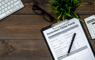 A clipboard with an insurance claim form, black pen, calculator, eyeglasses, keyboard, and potted plant on a wooden desk.