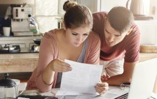 A woman and a man look at a document together at a kitchen table with a laptop, coffee pot, and papers in front of them.