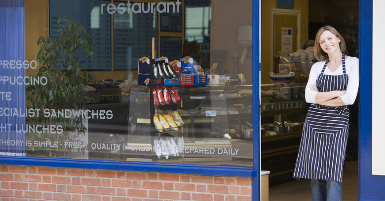 A woman wearing an apron stands and smiles outside the entrance of a small restaurant with large windows displaying food items inside.