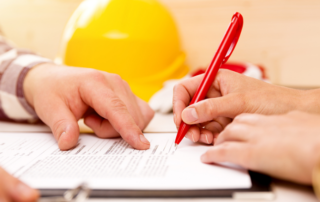 A person points to a document while another person signs it with a red pen; a yellow construction helmet is visible in the background.