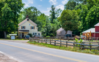 A rural roadside scene with a yellow house, a red barn, wooden fences, parked vehicles, and trees in the background on a sunny day.