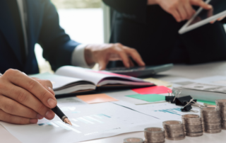 Two people working at a desk with financial documents, stacked coins, colorful sticky notes, an open notebook, and a tablet.
