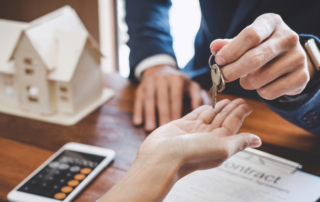 A person in a suit hands house keys to another person over a desk with a contract, smartphone, calculator, and a model house.