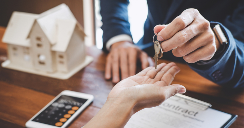 A person in a suit hands house keys to another person over a desk with a contract, smartphone, calculator, and a model house.