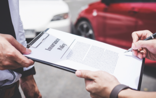 Two people review an insurance policy document on a clipboard, with a red car and a white car visible in the background.