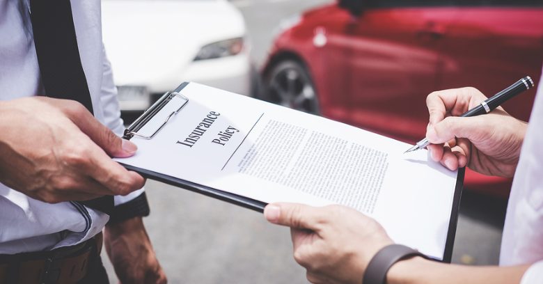 Two people review an insurance policy document on a clipboard, with a red car and a white car visible in the background.