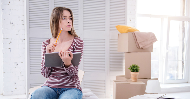 Woman holding a notebook and pencil sits on a bed, appearing thoughtful, with moving boxes and a plant visible in the background.