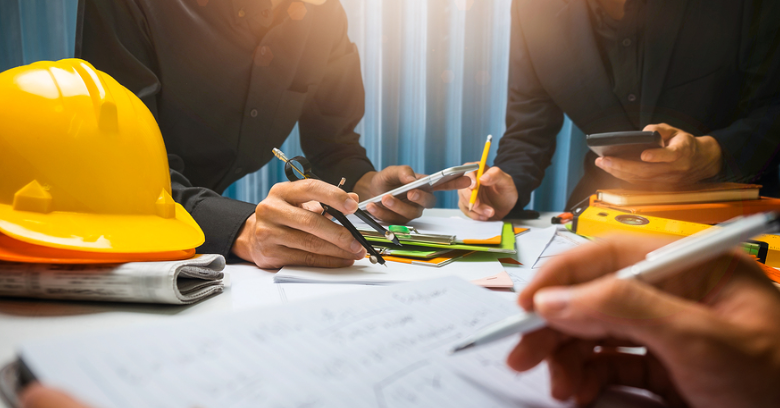 Three people work at a desk covered with papers, notebooks, and a yellow hard hat, reviewing documents and using a smartphone.