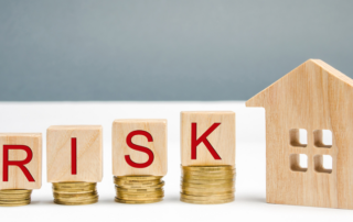 Wooden blocks spelling RISK are placed on stacks of coins next to a wooden house model, symbolizing financial risk in real estate or housing investments.