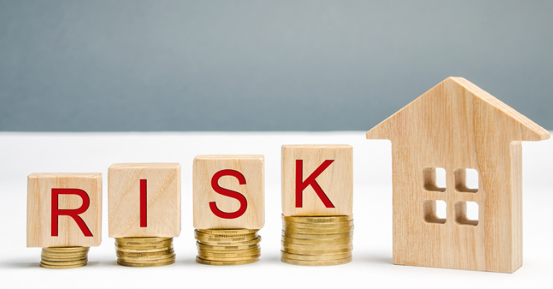 Wooden blocks spelling RISK are placed on stacks of coins next to a wooden house model, symbolizing financial risk in real estate or housing investments.