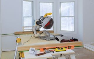 A miter saw is set up on a workbench in a room under construction, with tools, wood, and sawdust visible near three large windows.