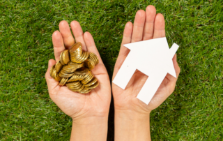 Two hands over grass; one holds a pile of coins, and the other holds a white paper cutout of a house.