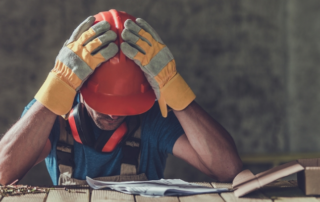 A construction worker wearing a hard hat and gloves sits at a table, holding his head in his hands and looking down at papers, appearing stressed or frustrated.