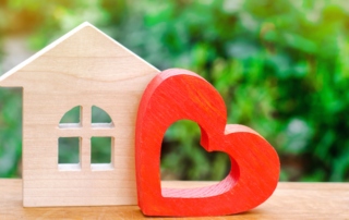 A small wooden house and a red heart-shaped figure are placed side by side on a wooden surface with greenery in the background.