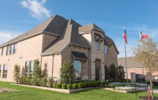 Two-story suburban house with brick exterior, manicured lawn, and landscaped garden beds. American and Texas flags are displayed on flagpoles near the entrance.