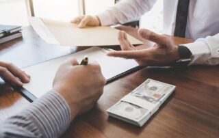 Two people at a desk; one hands over a folder labeled Contract while the other signs a document. A stack of hundred-dollar bills is on the table.