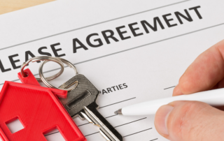 A person signs a lease agreement form next to a red house keychain and a set of keys.