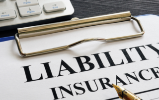 A clipboard holds a document labeled Liability Insurance next to a gold pen and part of a computer keyboard on a dark table.