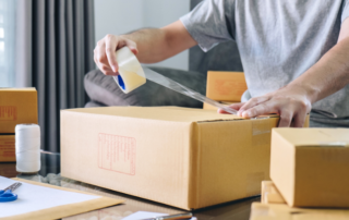Person sealing a cardboard box with clear packing tape on a table, surrounded by other boxes, scissors, and packing materials.