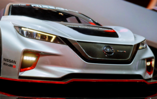 A silver Nissan Nismo concept car is displayed indoors, featuring sharp headlights, a mesh front grille, and red accents on the bumper.
