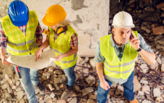 Three construction workers wearing safety gear stand among rubble; two review blueprints while one looks up and speaks into a radio.