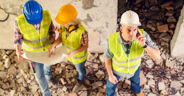 Three construction workers wearing safety gear stand among rubble; two review blueprints while one looks up and speaks into a radio.