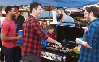 A group of people stand around a grill cooking burgers and hot dogs at an outdoor gathering, with some holding drinks and chatting.