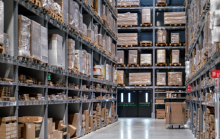 Large warehouse aisle with tall shelves holding neatly stacked boxes and pallets, illuminated by overhead lights, with double doors visible at the far end.