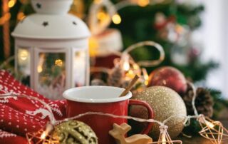 A red mug sits on a table with a wooden stir stick, surrounded by Christmas decorations, a white lantern, string lights, and a red patterned cloth.