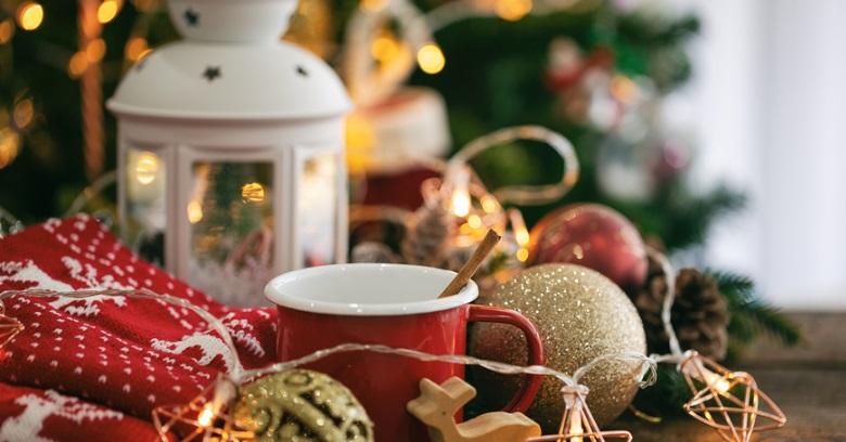 A red mug sits on a table with a wooden stir stick, surrounded by Christmas decorations, a white lantern, string lights, and a red patterned cloth.