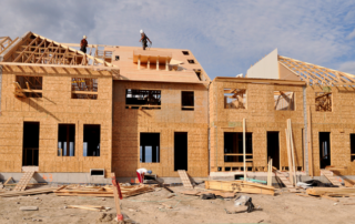 Partially constructed multi-unit building with exposed wooden framework; workers are assembling the roof under a partly cloudy sky.