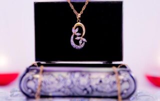 A gold necklace with a jeweled floral pendant is displayed on a decorative jewelry box with a black background and blurred lights in the background.