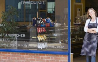 A woman wearing an apron stands smiling with arms crossed outside a cafe or restaurant, next to a large window displaying snacks and a menu board inside.