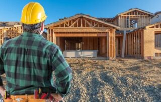 A construction worker in a hard hat and tool belt observes a partially built wooden house on a construction site.
