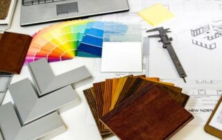 A workspace displays architectural plans, color swatches, wood and tile samples, a caliper, and a laptop, suggesting interior design or renovation planning.