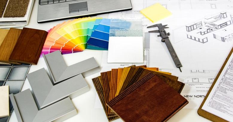 A workspace displays architectural plans, color swatches, wood and tile samples, a caliper, and a laptop, suggesting interior design or renovation planning.