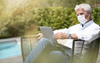 An older man wearing a mask sits outdoors by a pool, using a laptop and earphones, surrounded by greenery.