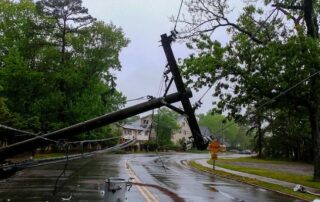 A utility pole is broken and leaning over a wet road, with downed power lines and debris visible; houses and trees are in the background on a cloudy day.